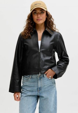 Woman with curly hair wearing a beige cap, black zip-up leather jacket, white top, and light blue jeans, standing with one hand in pocket.