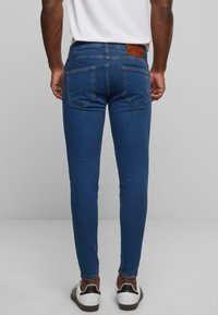Jeans de mezclilla azul ajustados con una textura suave, que presentan dos bolsillos traseros y una etiqueta de cuero marrón en la cintura, combinados con zapatillas blancas.
