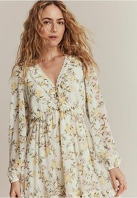 Blommig klänning i lätt tyg med v-ringning, lång ärm och volangdetaljer. Har gula och rosa blommor mot en vit bakgrund.