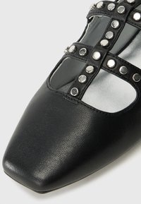 Zapato de cuero negro con puntera cuadrada, presenta cortes, remaches plateados y un interior metálico suave. Parte superior texturizada con acabado mate.