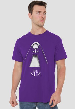 Man wearing purple T-shirt with graphic of a sinister nun face and text "The Nun," looking to the side against a plain light background.