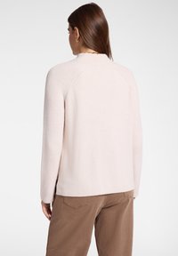Femme aux longs cheveux bruns portant un pull côtelé rose clair et un pantalon marron, debout de dos sur un fond blanc uni.