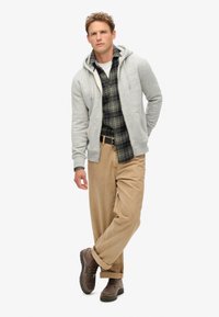 Grijze zip-up hoodie over een donkere geruite shirt, gecombineerd met beige broeken en bruine veterschoenen. Soepele textuur, casual pasvorm en gelaagde stijl.