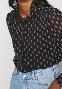 Sort bluse med små blomsterprint i rød og hvid, med en flæsekrave og knaplukning. Båret med lyseblå denimjeans.