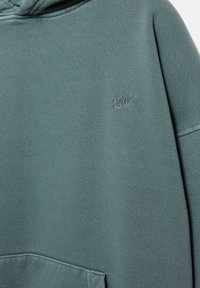 Turkizna kapuca z majhnim vezenim logotipom "SWT" na prsih in sprednjim žepom v slogu kenguruja, ki prikazuje teksturo tkanine in detajle šivov.