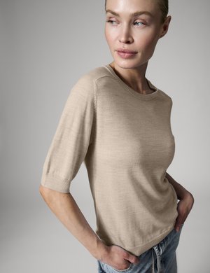 Femme portant un pull beige à manches courtes et un jean bleu, regardant sur le côté avec les mains dans les poches sur un fond gris uni.