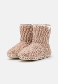 Knitted beige bootie slippers with a textured surface, rounded toe, and patterned white and brown sole detailing.
