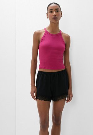 Femme debout contre un mur blanc portant un haut magenta sans manches et un short noir avec un bord en dentelle.