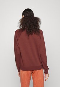 Mörkröd sweatshirt med raglanärmar, ribbad mudd och nederkant, samt en mjuk textur. Bärs med orange byxor som visar en avslappnad passform.