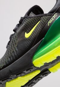 Svart atletisk sko med nätöverdel, ljusgula detaljer och en grön genomskinlig sula. Har strukturerad yta och förstärkt tå.