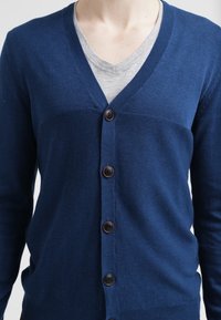 Mörkblå cardigan i stickad tyg, med v-ringad design, sex bruna knappar och en strukturerad passform. Ärmarna är långa och åtsittande.