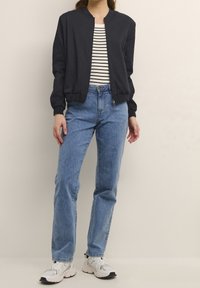 Giacca bomber nera con collo rotondo, abbinata a una maglietta a righe e jeans chiari a vita alta. Le sneaker presentano una combinazione di bianco e grigio.
