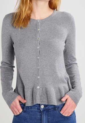 Kobieta w jasnoszarym, długim swetrze na guziki z falbanką u dołu i niebieskich dżinsach, ręce w kieszeniach.