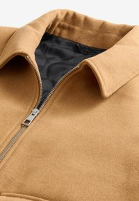 Chaqueta de lana color marrón claro con cuello doblado, cremallera plateada y forro acolchado negro. Exterior texturizado y detalles de diseño elegantes.