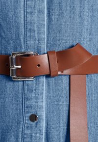 Brązowy skórzany pasek z metalową klamrą w kolorze srebrnym, przewleczony przez pętle na niebieskiej koszuli dżinsowej, z widocznym przeszyciem i zatrzaskiem.