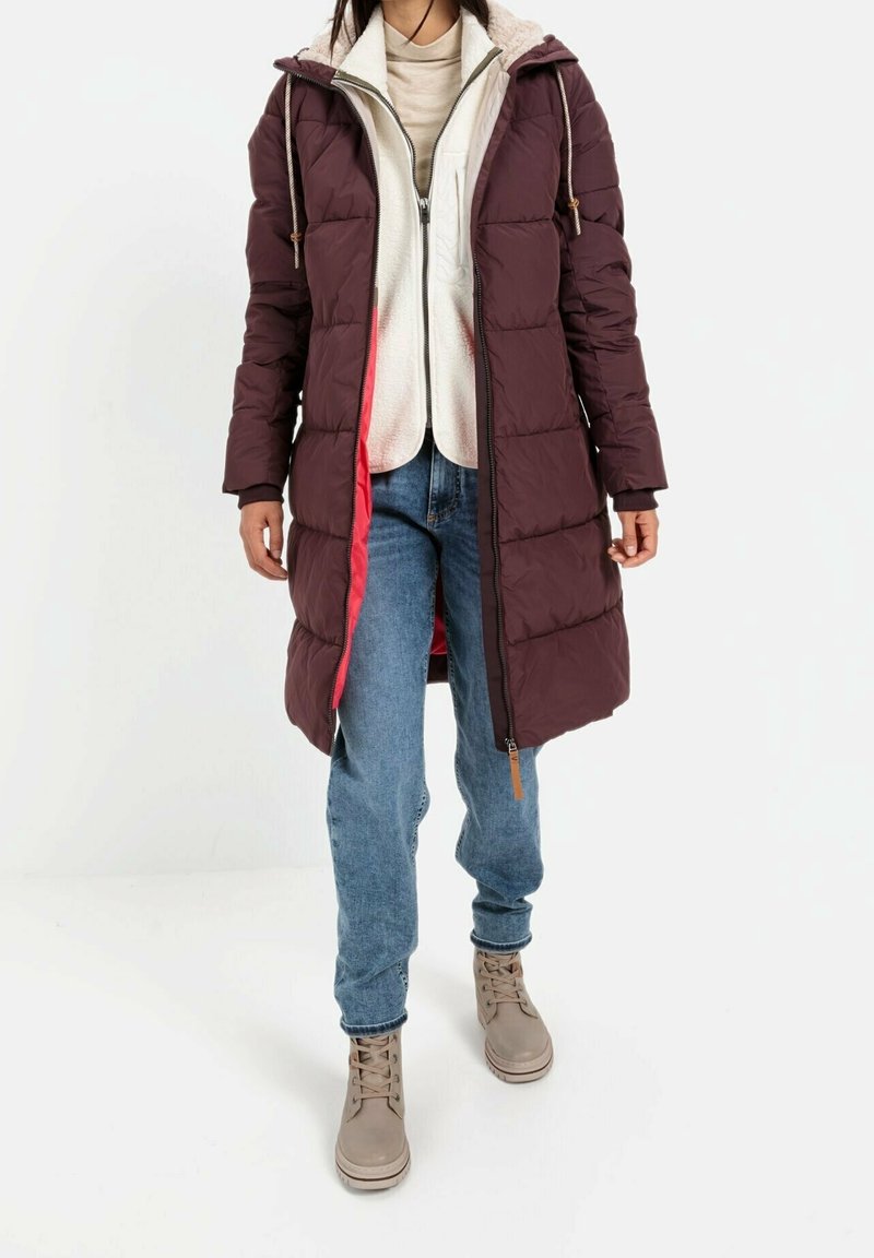 Manteau long matelassé bordeaux avec capuche, doté d'une doublure douce, fermeture éclair à l'avant et poches latérales, porté avec un jean bleu et des bottes beige.