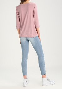 Helles Rosa, dreiviertel-langarm Oberteil mit lockerer Passform, kombiniert mit blauen Skinny-Jeans und weißen High-Top Sneakers. Schlichte, glatte Textur.