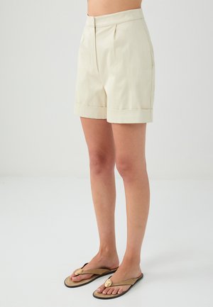 Knie-lange, lichtbeige op maat gemaakte shorts met plooien en opgevouwen manchetten, gemaakt van een soepele stof. Gekleed met bruine teenslippers met een gouden accent.