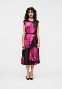 Robe noire avec un grand motif floral rose, sans manches, taille ceinturée et jupe fluide. Le mannequin porte des talons hauts noirs.