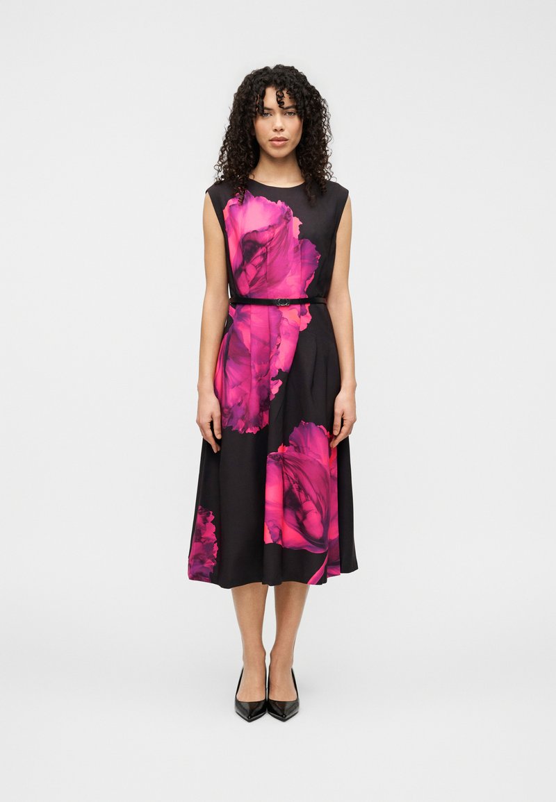 Robe noire avec un grand motif floral rose, sans manches, taille ceinturée et jupe fluide. Le mannequin porte des talons hauts noirs.