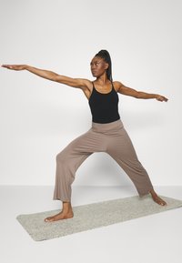 Black tank top and light brown wide-leg pants worn by a person in a yoga pose on a textured gray mat against a plain background.