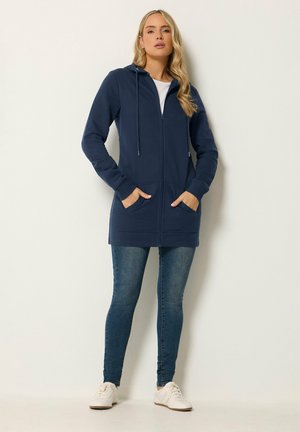 Vrouw staand, gekleed in een marineblauwe hoodie met rits en zakken, blauwe jeans en witte sneakers tegen een effen lichte achtergrond.