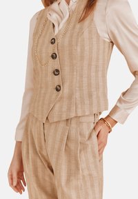 Beige gestreepte linnen vest met vijf donkere knopen, gecombineerd met bijpassende geplooide broeken en een lichtgekleurde overhemd eronder. Gouden ketting als accent.