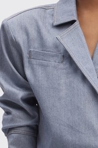 Ljusblå denimjacka med nothals, en bröstficka och framträdande axelsömmar. Har subtila sömmar och en mjuk textur.