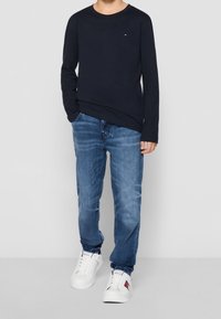 Marinblå långärmad skjorta, blå denimjeans och vita sneakers. Skjortan har en liten logotyp; jeansen har en klassisk passform med lätt slitage.