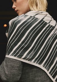 Leichter Schal aus halbtransparentem Stoff mit einem schwarz-weißen Streifenmuster, drapiert über einem strukturierten grauen Oberteil.