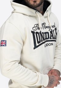 Sudadera crema con un logo negro que dice "The Original Lonsdale London". Cuenta con capucha con cordón y un parche de la bandera de la Unión en la manga.