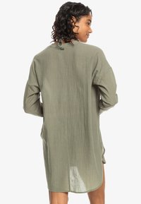Tunika verde oliva a maniche lunghe con vestibilità comoda, tessuto testurizzato e dettaglio sul retro con nodo. Le spaccature laterali migliorano la mobilità.