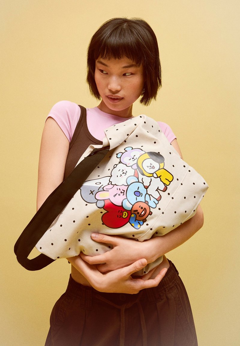 Femme tenant un sac cabas à pois avec des personnages de dessins animés colorés, portant un haut rose et un pantalon marron, debout sur un fond jaune.