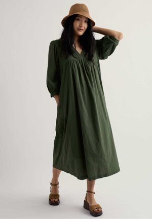 Woman wearing a loose olive green midi dress, brown platform sandals, and a woven brown bucket hat, posing with one hand on her head.