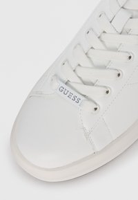 White sneaker made of smooth synthetic material, featuring round toe, flat white laces, and a logo tag labeled "GUESS" on the tongue.