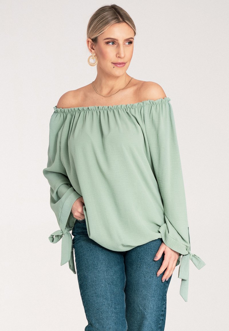 Jasnozielona bluzka z odkrytymi ramionami, z elastycznym dekoltem, długimi rękawami i wiązaniami przy mankietach. Materiał wydaje się miękki i lekko teksturowany.