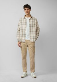 Koszula w kratkę w odcieniach beżu i brązu, biała koszulka, beżowe spodnie cargo z kieszeniami oraz jasne trampki z szarymi akcentami.