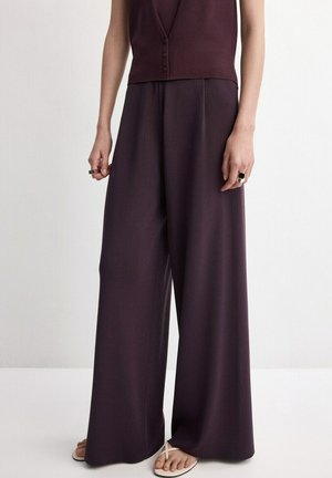 Mujer vistiendo pantalones anchos y sueltos de color púrpura oscuro, combinados con un top sin mangas abotonado y sandalias beige, posando contra un fondo liso.