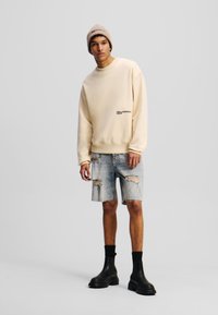 Kremasti sweatshirt s logotipom, uparen s iznošenim trapericama šortsevima. Nosivo s crnim gležnjačama i bež pletenom kapom.