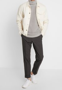 Mann in einer cremefarbenen Jacke über einem grauen Rollkragenpullover, dunklen grauen Hosen und weißen Sneakers, der mit den Händen in den Taschen steht.