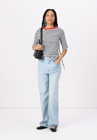 Gestreiftes schwarz-weißes T-Shirt mit rotem Ausschnitt, hellblaue weit geschnittene Jeans und schwarze flache Schuhe. Trägt eine kleine schwarze Tasche.