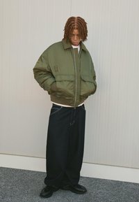 Giacca bomber verde con chiusura a zip, polsini a costine e due tasche frontali, abbinata a jeans in denim scuro a vestibilità ampia e scarpe nere.