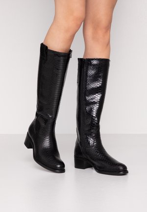 Czarne kozaki za kolano z błyszczącym, teksturowanym wzorem przypominającym skórę węża, noszone na gołych nogach, stojące na białej powierzchni na tle jednolitym.