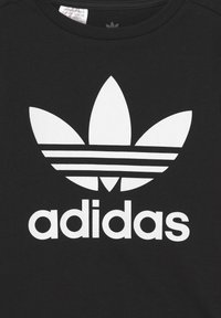 Črna majica s kratkimi rokavi, ki ima velik bel Adidasov trefoil logotip in ime blagovne znamke v odmevnih črkah, s krožnim izrezom in bombažno teksturo.