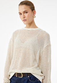 Sweter w kolorze kremowym, wykonany z luźno dzianej tkaniny, o swobodnym kroju, okrągłym dekolcie i długich rękawach. Połączony z ciemnym paskiem i niebieskimi dżinsami.
