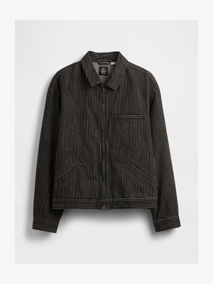 Chaqueta negra de cremallera con rayas verticales blancas, que cuenta con un cuello, dos bolsillos laterales y un bolsillo de ojal en el pecho.