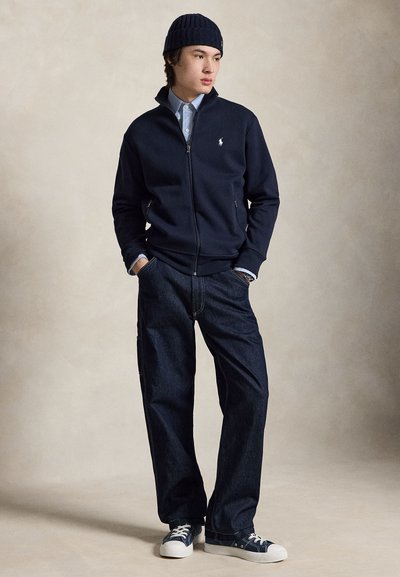 Young man wearing a navy zip-up jacket, blue button-up shirt, dark jeans, navy beanie, and blue sneakers with white soles and laces.