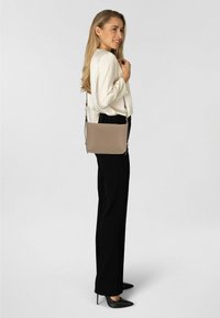 Sac bandoulière beige avec une finition lisse, une sangle réglable et des éléments en métal argenté. Porté par une personne vêtue d'un chemisier blanc et d'un pantalon noir.