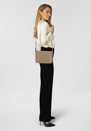 Bolso bandolera beige con acabado liso, correa ajustable y herrajes plateados. Llevado por una persona con blusa blanca y pantalones negros.
