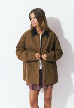 Veste en coton marron avec un col foncé, des boutons-pression à l'avant et de grandes poches. Portée par-dessus une chemise blanche et un short violet. Texture lisse.